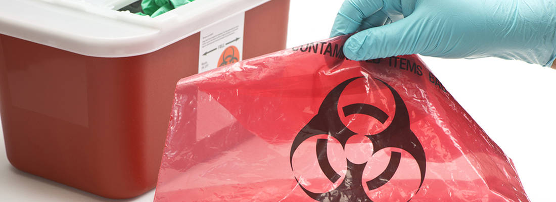 Gloved hand holds Red bag with hazardous waste symbol. Red sharps box sits behind 
