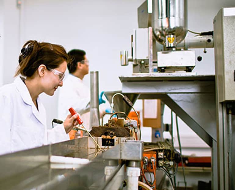 Two engineers in white lab coats and safety glasses work on a production line 