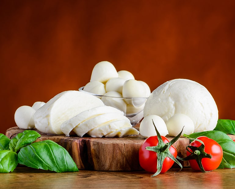 Mozzarella cheese sits on a wooden chopping board with herbs and tomatoes in front 