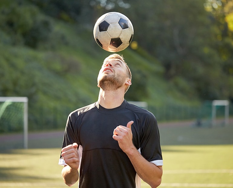 Male in black t-shirt heading a football on football pitch