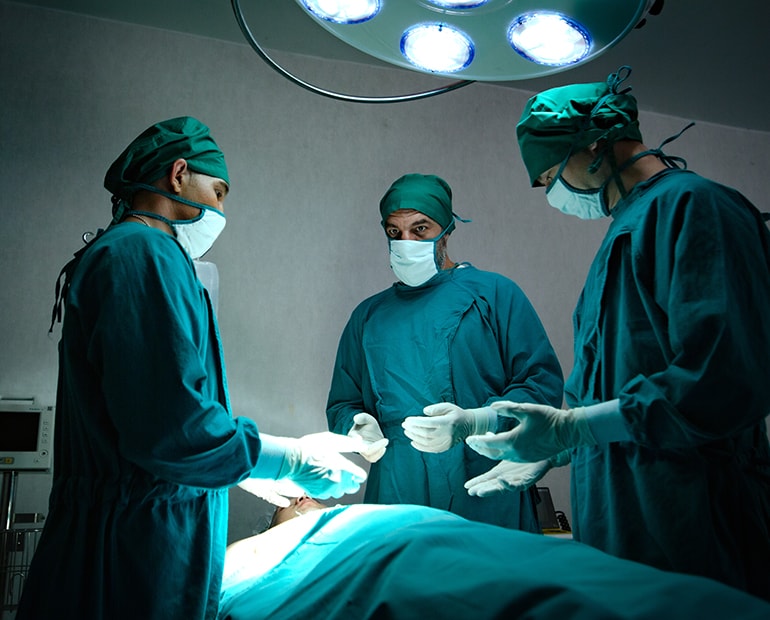 The surgeons with green gown, gloves, mask and cap stand over an operating table 