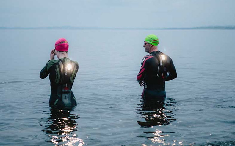 Female and male swimmers, waist deep in the sea, wearing wetsuits with a Tekralite on their backs.
