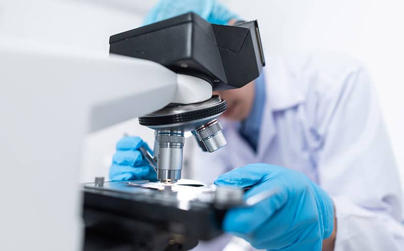 Male researcher wearing a white lab coat and blue gloves inspects a slide on a microscope