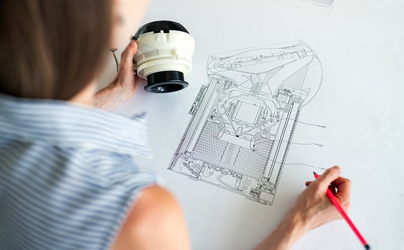 Female engineer looking at technical drawing of prototype, whilst holding a prototype in left hand