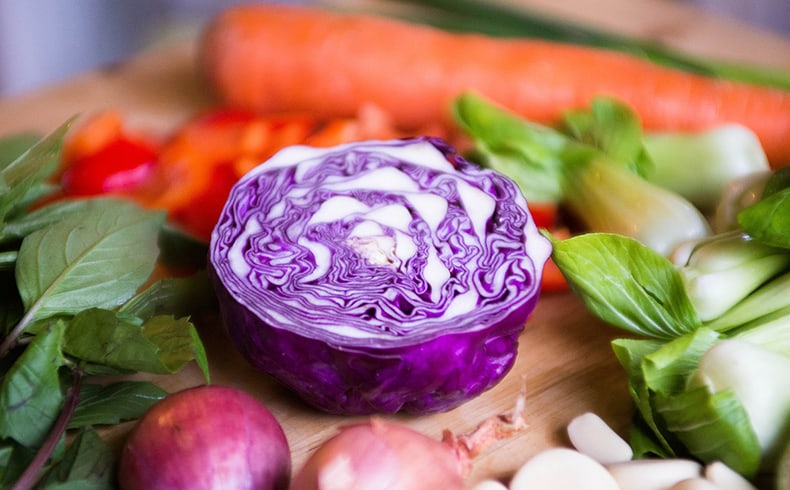 Cut red cabbage sits on a wooden chopping board surrounded by a selection of vegetables