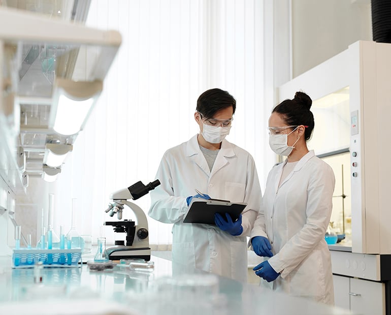 Male and Female researchers wearing white lab coats in a lab. Male researcher holds a clipboard 