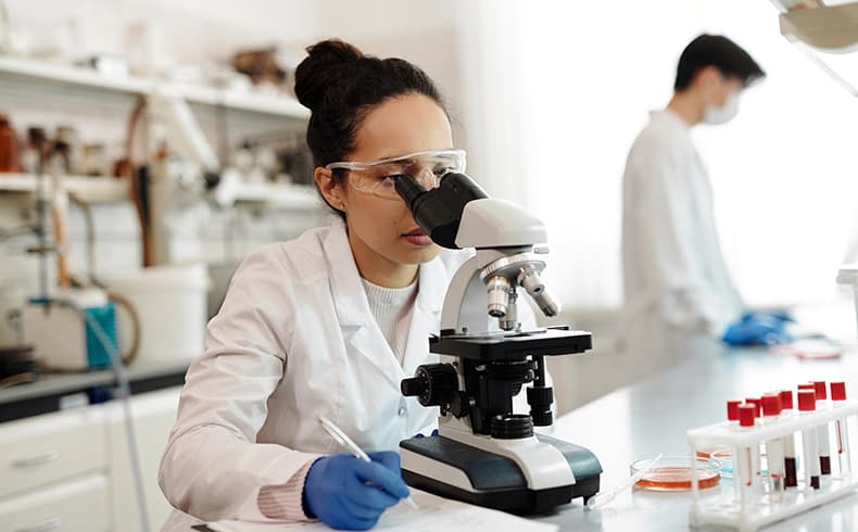 Female researcher in a lab wearing a white lab coat and safety glasses looking through a microscope. Male researcher in white lab coat in the background.